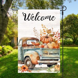 Könnte beinhalten: Eine Willkommens-Gartenflagge mit einem blauen Vintage-Pickup, der einen großen Haufen Kürbisse transportiert. Der LKW hat die Aufschrift "The Smith's" auf der Rückseite. Die Flagge hat einen weißen Holzmaserungshintergrund mit dem Wort "Welcome" in schwarzer Kursivschrift.