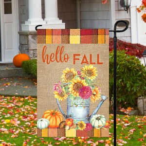 May include: A burlap garden flag with a plaid border and the text "hello FALL". The flag features a watercolor illustration of sunflowers and other fall flowers in a blue watering can with pumpkins and gourds at the bottom.