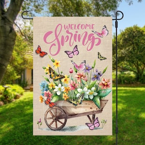May include: A garden flag with a burlap background featuring a wheelbarrow overflowing with colorful flowers and butterflies. The text "Welcome Spring" is written in pink cursive lettering.