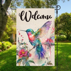 Può includere: Una bandiera da giardino di benvenuto con un colibrì colorato in volo, circondato da fiori rosa e schizzi in stile acquerello. La bandiera ha la parola "Benvenuto" stampata in lettere corsive nere.