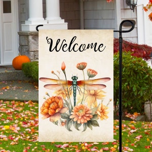 Puede incluir: Una bandera de jardín de bienvenida con un diseño de libélula y flores. La bandera es beige con un fondo marrón y presenta una libélula con alas naranjas y cuerpo verde. La libélula está rodeada de flores naranjas y amarillas.