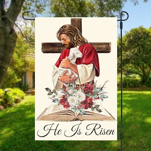 Pode incluir: Uma bandeira de jardim com fundo branco que apresenta uma pintura de Jesus segurando um cordeiro. Ele está de pé em frente a uma cruz de madeira com uma túnica vermelha e uma roupa interior branca. Um livro com rosas vermelhas e flores brancas está na parte inferior da imagem. O texto "He Is Risen" está escrito em itálico na parte inferior da bandeira.