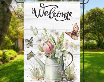 May include: A white garden flag with a wooden plank background featuring a watercolour illustration of a watering can filled with pink and white flowers, green leaves, and butterflies. The text "Welcome" is written in black cursive lettering at the top of the flag.