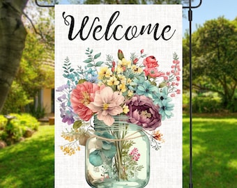 May include: A welcome garden flag with a white background featuring a mason jar filled with a colourful bouquet of flowers. The text "Welcome" is written in black cursive at the top of the flag.