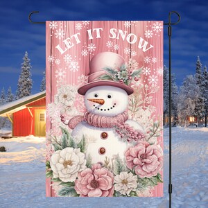 May include: A pink and white snowman with a pink hat and scarf surrounded by pink and white flowers. The background is a pink wooden fence with the text "Let It Snow" in white.