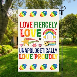 Puede incluir: Una bandera de jardín con las palabras "LOVE FIERCELY LOVE UNAPOLOGETICALLY LOVE PROUDLY" en varios colores. La bandera presenta labios de arcoíris, un arcoíris, estrellas y flores. La bandera se muestra al aire libre en un jardín.