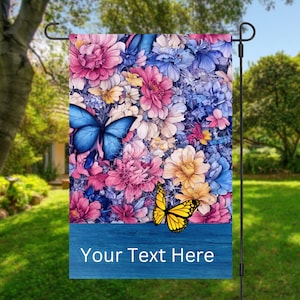 May include: A garden flag with a blue background and a floral design featuring pink, yellow, and blue flowers. Two butterflies, one blue and one yellow, are featured on the flag. The text "Your Text Here" is printed on the bottom of the flag.