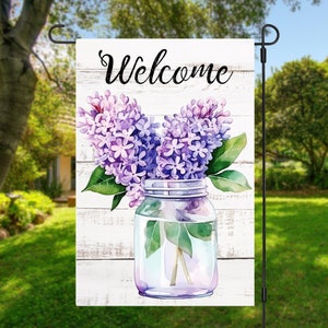 Peut inclure: Une pancarte en bois blanc avec le mot "Bienvenue" en écriture noire. Un bocal en verre rempli de fleurs de lilas violettes est placé devant la pancarte.