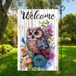 May include: A white wooden sign with the word "Welcome" in black script. A brown owl with yellow eyes is perched on a bed of colourful flowers, including pink roses, purple flowers, and blue flowers.