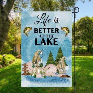 Könnte beinhalten: Eine blaue und weiße Gartenflagge mit zwei Gnomen, die angeln. Die Flagge trägt den Schriftzug "Life is Better at the Lake" mit zwei Fischen auf jeder Seite des Textes.