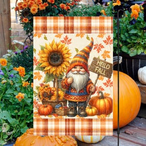 Könnte beinhalten: Eine herbstliche Gartenflagge mit einem Zwerg, der ein "Hello Fall" Schild hält, umgeben von Sonnenblumen, Kürbissen und Herbstblättern. Die Flagge hat einen karierten Rand und einen cremefarbenen Hintergrund.