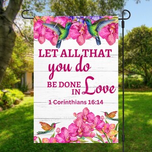 May include: A white wooden garden flag with pink orchids and hummingbirds. The flag has the text "Let all that you do be done in love 1 Corinthians 16:14".