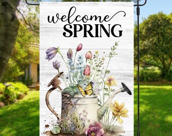 May include: A white wooden garden flag with the text "welcome SPRING" in black lettering. A watercolour illustration of a watering can filled with colourful flowers and a yellow butterfly is in the centre of the flag.