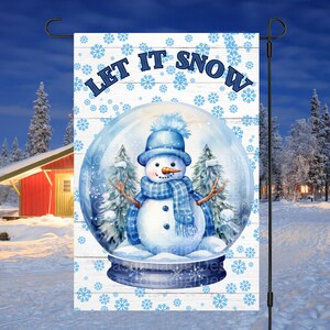 May include: A white garden flag with a blue and white snowman inside a snow globe. The snow globe is on a white wooden background with a blue snowflake pattern. The text "Let It Snow" is written in blue letters at the top of the flag.