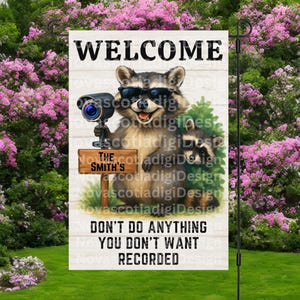 Puede incluir: Una bandera de jardín con un mapache con gafas de sol y sosteniendo una cámara de seguridad. La bandera dice "WELCOME" en la parte superior y "DON'T DO ANYTHING YOU DON'T WANT RECORDED" en la parte inferior. Un letrero dice "THE SMITH'S".