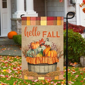May include: A burlap garden flag with a plaid border and the text "hello FALL" in orange. A wooden bucket filled with pumpkins, gourds, and fall foliage is in the center of the flag.