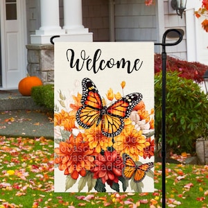 Könnte beinhalten: Eine Willkommens-Gartenflagge mit weißem Hintergrund, auf der ein großer orangefarbener und schwarzer Schmetterling mit orangefarbenen und roten Blumen abgebildet ist. Der Text "Willkommen" ist in schwarzer Kursivschrift geschrieben.