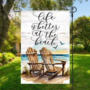 May include: A garden flag with a white wooden background and the text "Life is better at the beach". Two brown wooden chairs are on a sandy beach with a blue ocean and a sunset in the background.