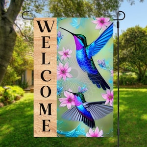 May include: A welcome garden flag with the word "WELCOME" printed on a wooden background. The flag features two colorful hummingbirds flying among pink flowers on a green background.