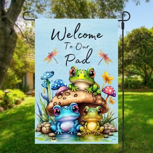 May include: A blue and white garden flag with the text "Welcome to Our Pad" and three cartoon frogs sitting on a mushroom. The frogs are green, blue, and yellow.