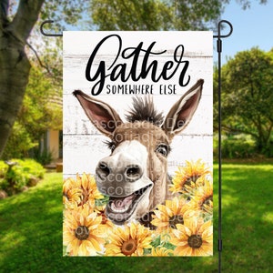 May include: A garden flag with a white wooden background and the text "Gather Somewhere Else". A brown donkey with a big smile is peeking out from behind a bunch of yellow sunflowers.