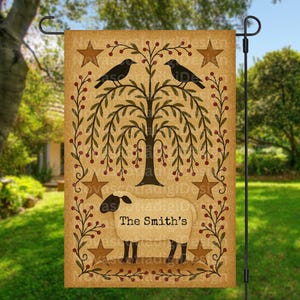 Könnte beinhalten: Eine beige Gartenflagge mit einem Baum, zwei schwarzen Vögeln, Sternen und einem Schaf. Der Text "The Smith's" befindet sich unter dem Schaf. Die Flagge wird auf einem schwarzen Metallständer präsentiert, ideal für die Außendekoration.