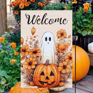 以下が含まれることがあります： 黒い筆記体で「Welcome」と書かれたハロウィーンのガーデンフラッグ。旗には、笑顔の幽霊、オレンジ色の花、ジャック・オー・ランタンが描かれています。背景はベージュ色です。