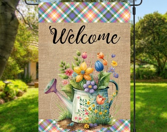 May include: A garden flag with a burlap background and a colourful plaid top and bottom border. The flag features a watering can overflowing with flowers and the word "Welcome" in elegant script.