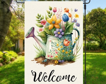 May include: A welcome garden flag featuring a watercolour illustration of a blue watering can overflowing with colourful flowers. The text "Welcome" is printed in black at the bottom of the flag.