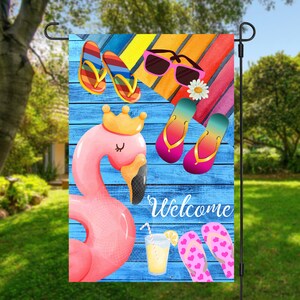 May include: A summer-themed welcome garden flag featuring a pink flamingo, sunglasses, flip-flops, and a glass of lemonade on a blue wooden background. The text "Welcome" is written in white cursive.