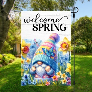 May include: A welcome spring garden flag with a gnome wearing a colorful hat and a butterfly on top. The gnome is surrounded by yellow daffodils and blue flowers. The flag has a white wooden background with the text "welcome spring" in black.