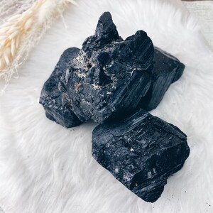 Puede incluir: Un conjunto de cristales de turmalina negra en bruto. Los cristales tienen una superficie rugosa e irregular y tama&ntilde;os variables. Se disponen sobre una superficie blanca y esponjosa, con un tallo de planta seca en el fondo.