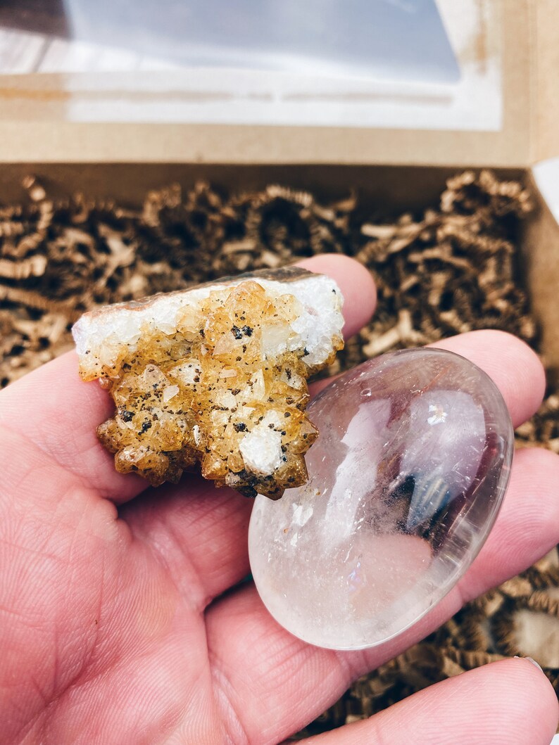 Puede incluir: Un c&uacute;mulo de peque&ntilde;os cristales de citrino amarillos con un cristal de cuarzo redondo transparente. Ambos cristales se sostienen en una mano.