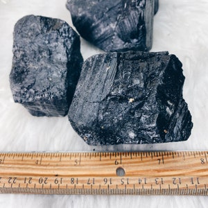 Puede incluir: Tres piedras de turmalina negra en bruto con una regla para la escala. Las piedras tienen una superficie texturizada e irregular y var&iacute;an en tama&ntilde;o. La regla muestra medidas en cent&iacute;metros, con la piedra m&aacute;s grande midiendo aproximadamente 15 cm de largo.