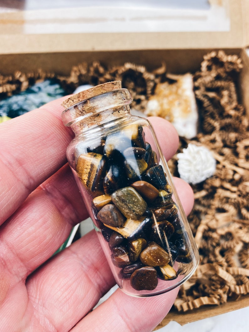 Puede incluir: Una peque&ntilde;a botella de vidrio llena de piedras de ojo de tigre. Las piedras son una mezcla de marr&oacute;n, dorado y negro.