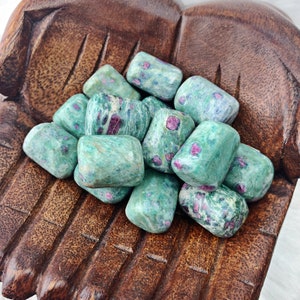 May include: A collection of green and red tumbled stones with a speckled pattern. The stones are arranged in a wooden hand-shaped bowl.