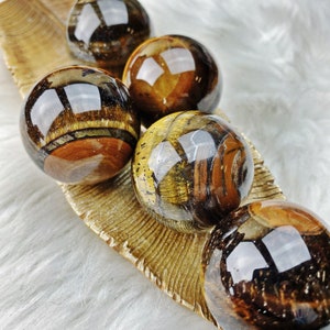 May include: Five polished tiger's eye spheres in various shades of brown, gold, and black. The spheres are arranged on a gold-colored leaf-shaped surface.