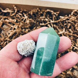 Puede incluir: Una punta de cristal verde y un cristal de cuarzo rosa blanco. Los cristales est&aacute;n en una mano, con trozos de papel marr&oacute;n en el fondo.