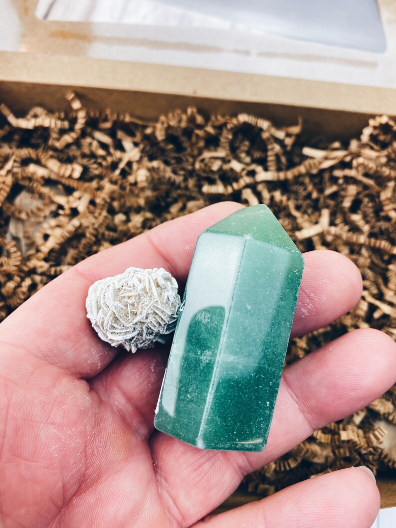Puede incluir: Una punta de cristal verde y un cristal de cuarzo rosa blanco. Los cristales est&aacute;n en una mano, con trozos de papel marr&oacute;n en el fondo.
