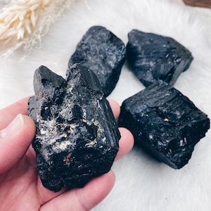 Puede incluir: Cuatro cristales de turmalina negra en bruto. Los cristales son de color negro intenso con superficies texturizadas. Un cristal se sostiene en una mano, mientras que los otros est&aacute;n dispuestos sobre una superficie blanca y esponjosa. Los cristales son un producto natural y terroso.
