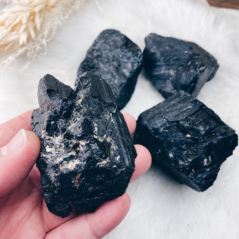 Puede incluir: Cuatro cristales de turmalina negra en bruto. Los cristales son de color negro intenso con superficies texturizadas. Un cristal se sostiene en una mano, mientras que los otros est&aacute;n dispuestos sobre una superficie blanca y esponjosa. Los cristales son un producto natural y terroso.