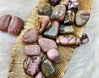 May include: A collection of pink and black tumbled stones, known as Rhodonite, on a gold leaf background. The stones are smooth and polished, with a variety of shapes and sizes.