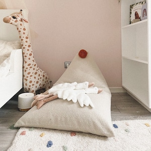 Puede incluir: Un puf blanco con un pompón en la parte superior. Dos cisnes de peluche blancos están tumbados en el puf. Un animal de peluche en forma de jirafa está de pie junto al puf. Una estantería blanca está en el fondo.