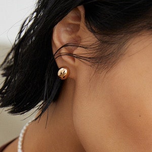 Puede incluir: Primer plano de una mujer con una camisa de manga larga marrón, un collar de perlas y pendientes de oro. La camisa tiene un escote en V profundo.