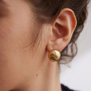 Puede incluir: Un par de pendientes de botón redondos martillados de color dorado. Los pendientes los lleva una persona con el pelo oscuro y un collar de oro.