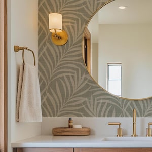 May include: A bathroom scene featuring a round gold-framed mirror, a gold faucet, and a matching towel ring. The wall is adorned with a green and white leaf pattern wallpaper. A beige towel hangs on the ring, and a small wooden tray holds toiletries.