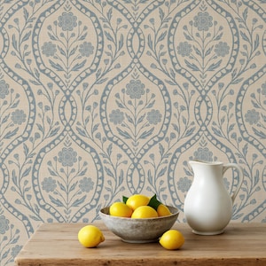 May include: A still life featuring a wooden table with a bowl of lemons, a white pitcher, and a floral patterned wallpaper in the background. The wallpaper has a light beige background with a blue floral design. The lemons are bright yellow.