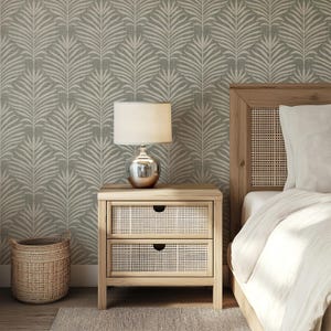 May include: A wooden nightstand with two drawers and a woven rattan front. The nightstand is next to a bed with a white duvet and a woven headboard. A silver lamp with a white shade sits on top of the nightstand. The wall behind the bed is covered in a green and white wallpaper with a palm leaf pattern.