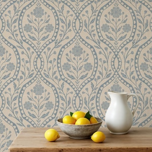 May include: A wooden table holds a gray bowl filled with yellow lemons, a white ceramic pitcher, and two loose lemons. The background features wallpaper with a repeating blue floral damask pattern on a light beige textured surface.