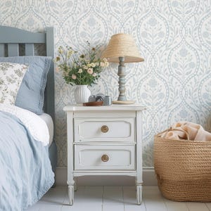 May include: A white bedside table with two drawers and brass knobs. A lamp with a straw shade sits on top, next to a vase of flowers and a vintage camera. A woven basket is on the floor.